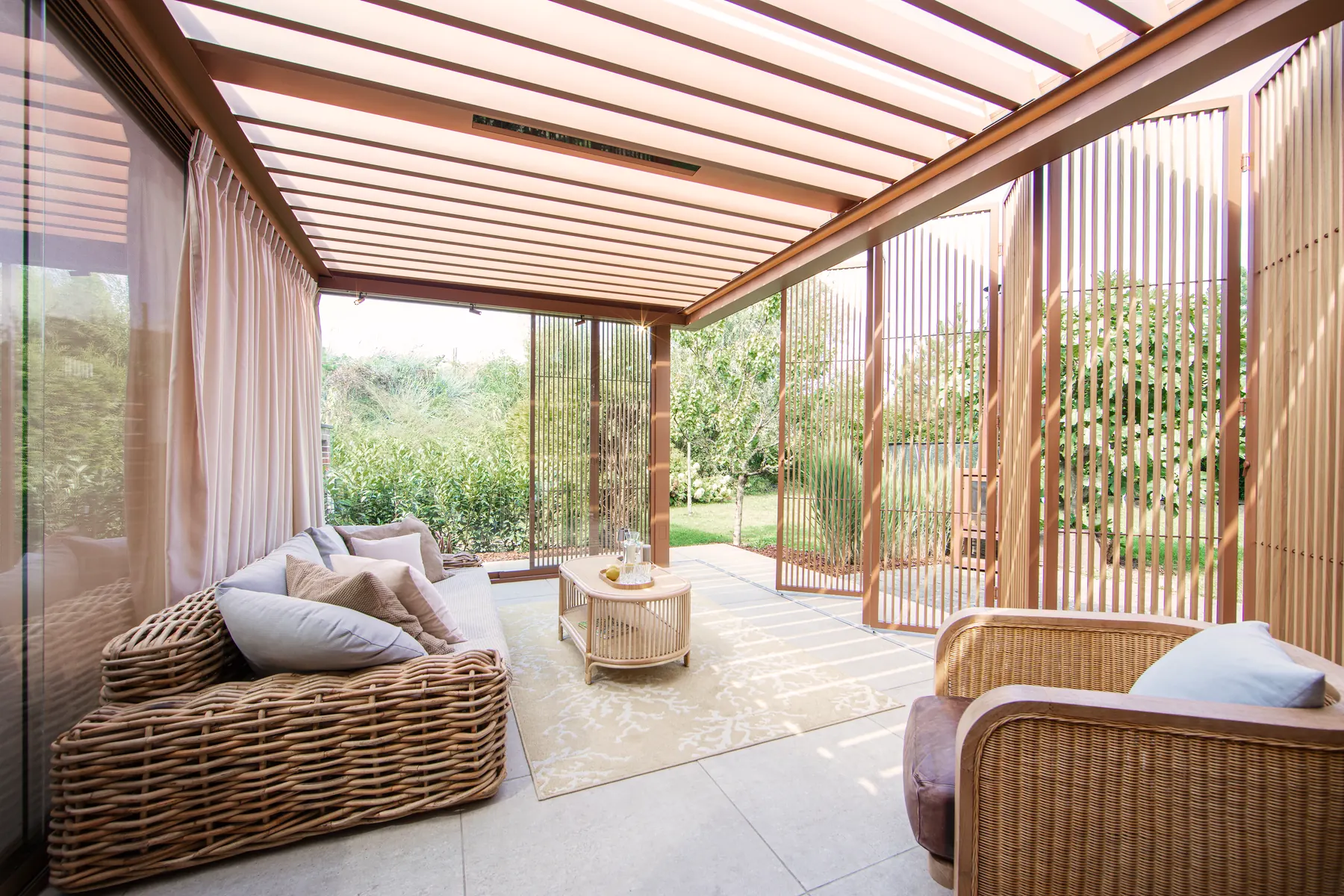 Lounge mit Korbmöbeln unter geöffneten Lamellen, Vorhänge und seitliche Holzlamellen, Blick in den Garten