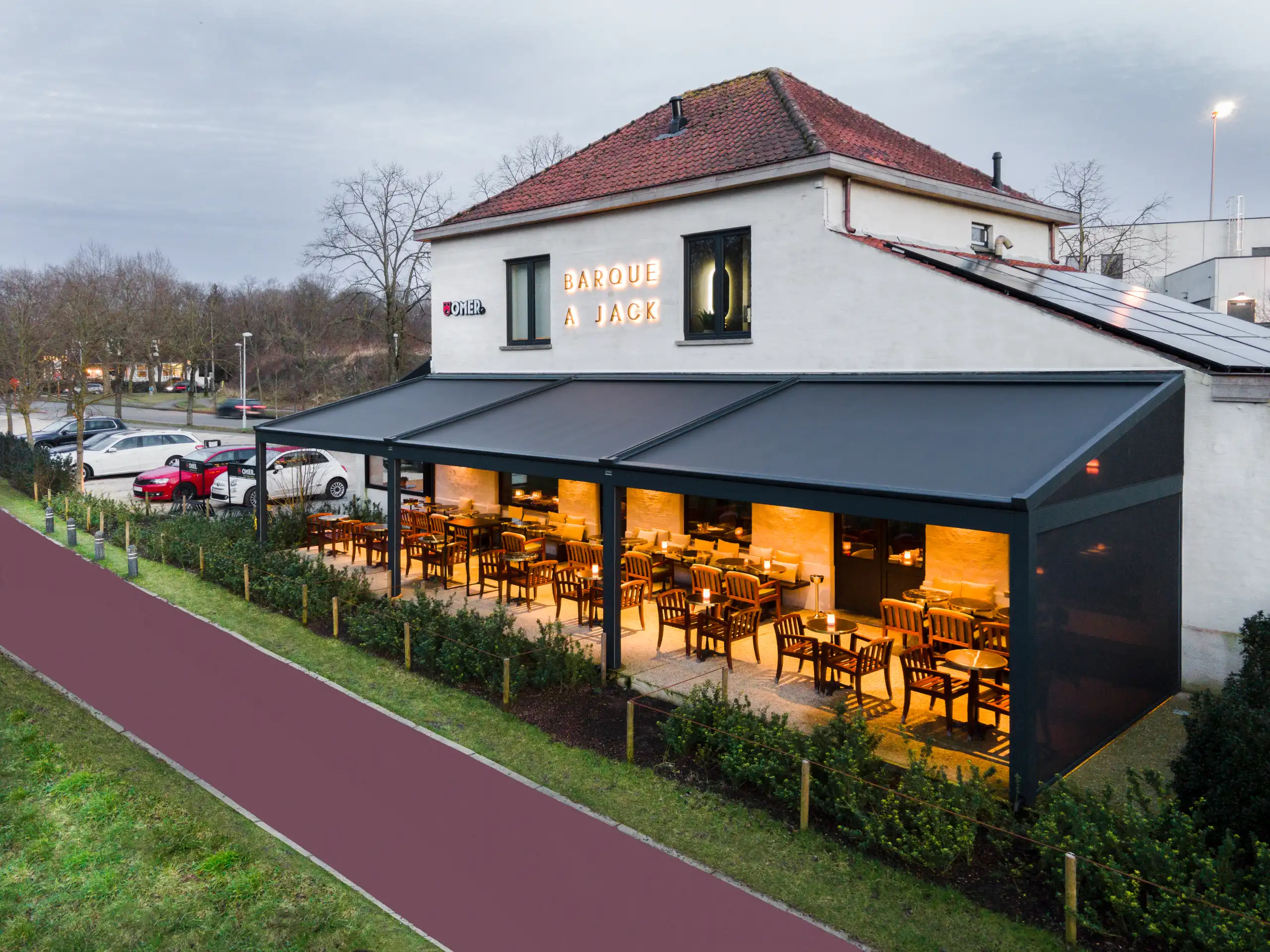 Luftaufnahme einer Restaurantterrasse in der Dämmerung: Das Gebäude hat eine dunkle Markisenüberdachung, darunter warm beleuchtete Tische und Stühle