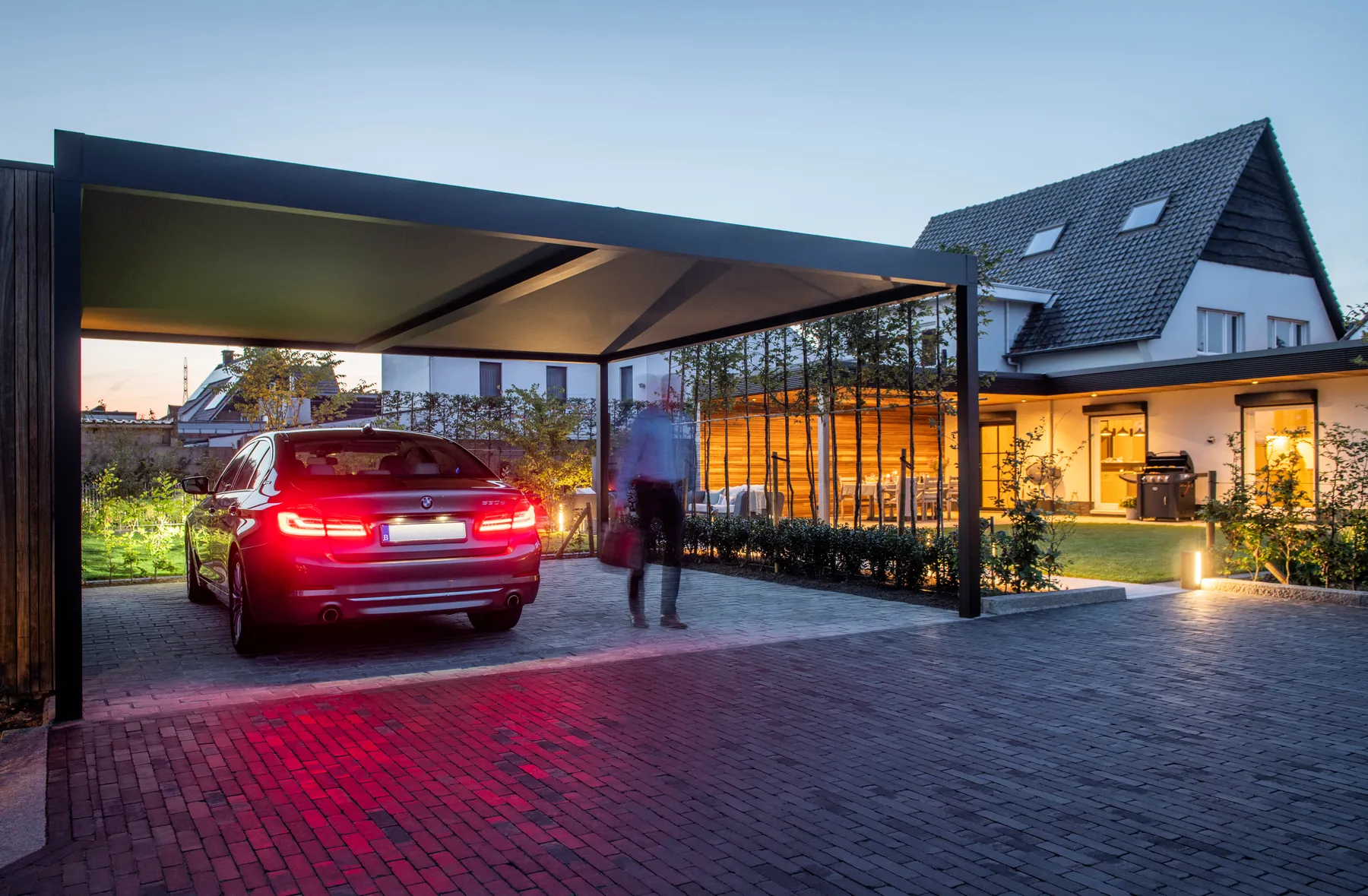 Carport in der Dämmerung, darunter ein Auto mit leuchtenden Rückleuchten, dahinter die beleuchtete Terrasse eines Hauses