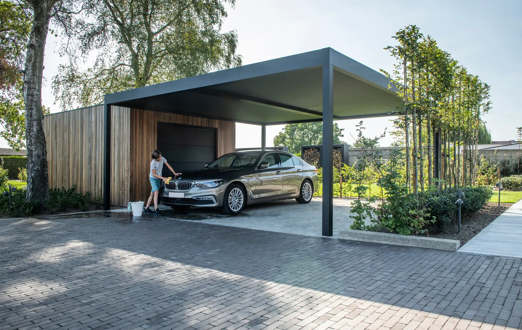 Carport mit dunklem Dach neben einem Holzhaus, darunter wird ein Auto von Hand gewaschen, im Hintergrund Bäume und Garten