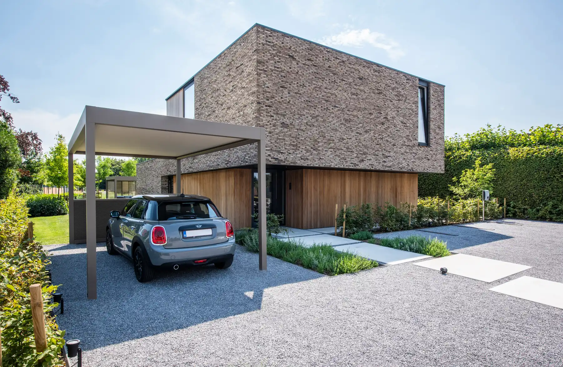 Freistehender Carport mit hellem Dach vor einem modernen Backsteinhaus, darunter ein kleiner grauer Wagen auf Kies