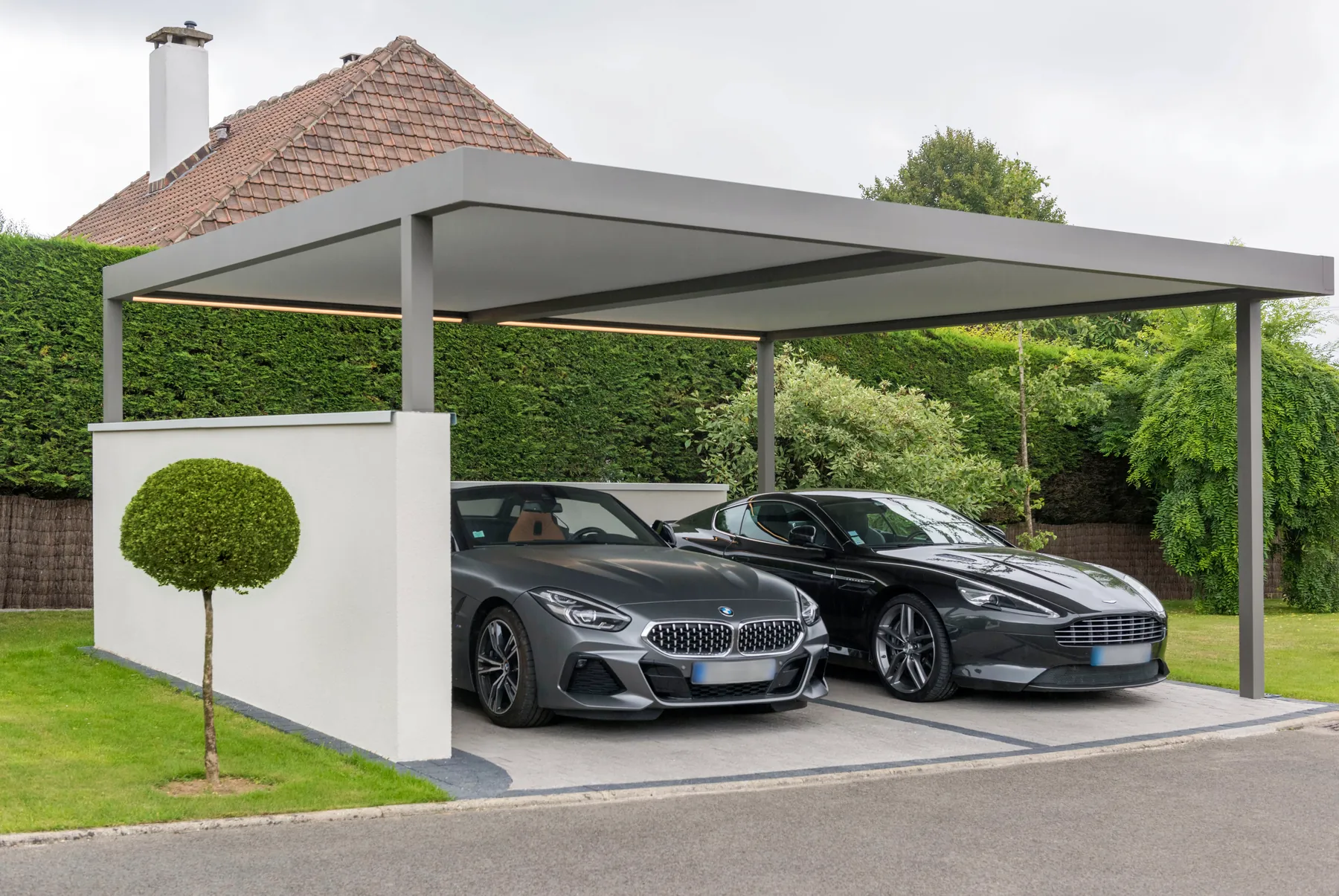 Doppelcarport mit hellgrauem Dach und weißer Wand, darunter zwei Sportwagen, davor ein kugelförmig geschnittener Baum