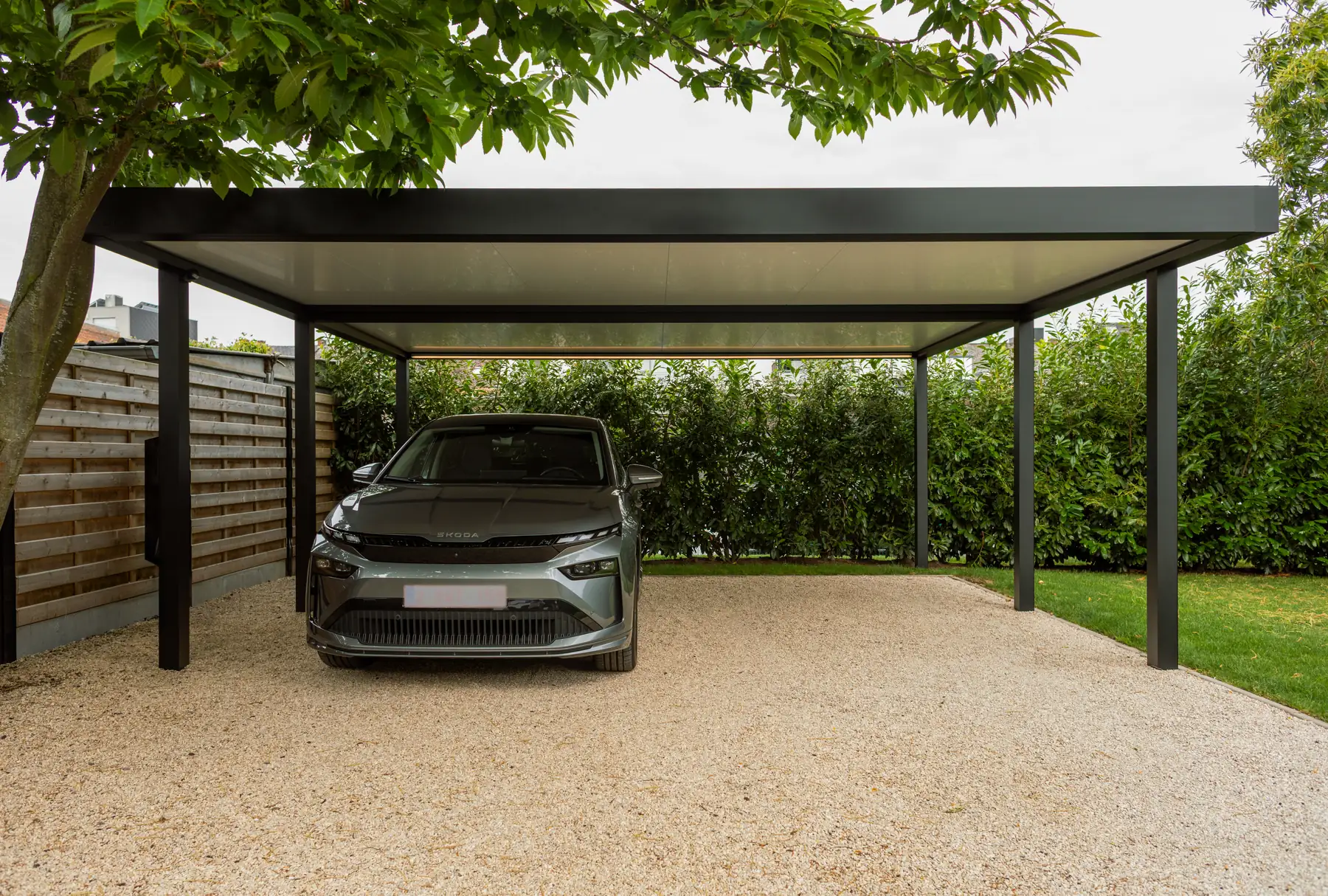 Großer Carport mit dunklem Dach über einem Elektroauto in Frontansicht auf hellem Kies, im Vordergrund ein Laubbaum
