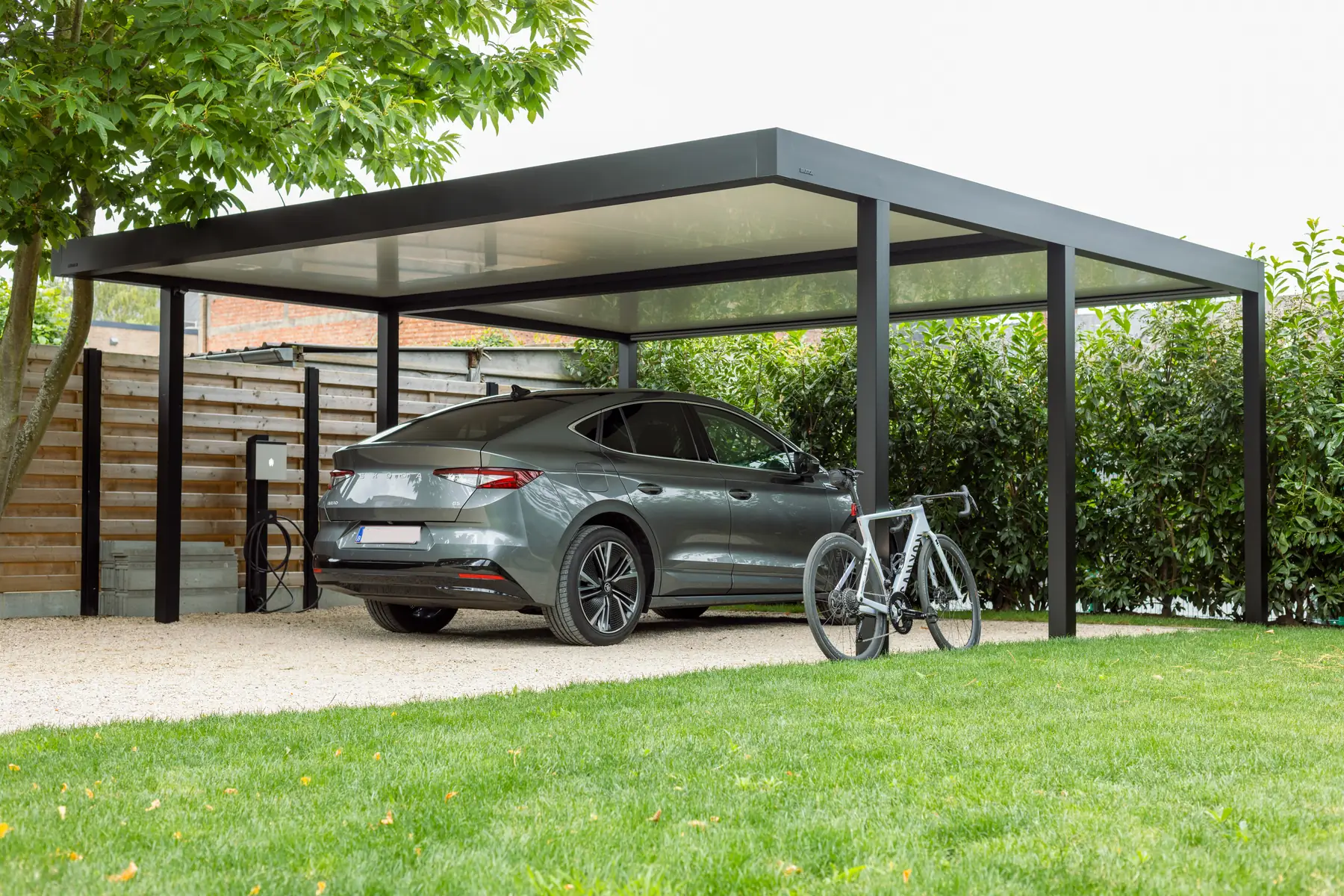 Carport mit dunklen Pfosten über einem Elektroauto an der Ladestation, davor ein Rennrad, dahinter eine Hecke