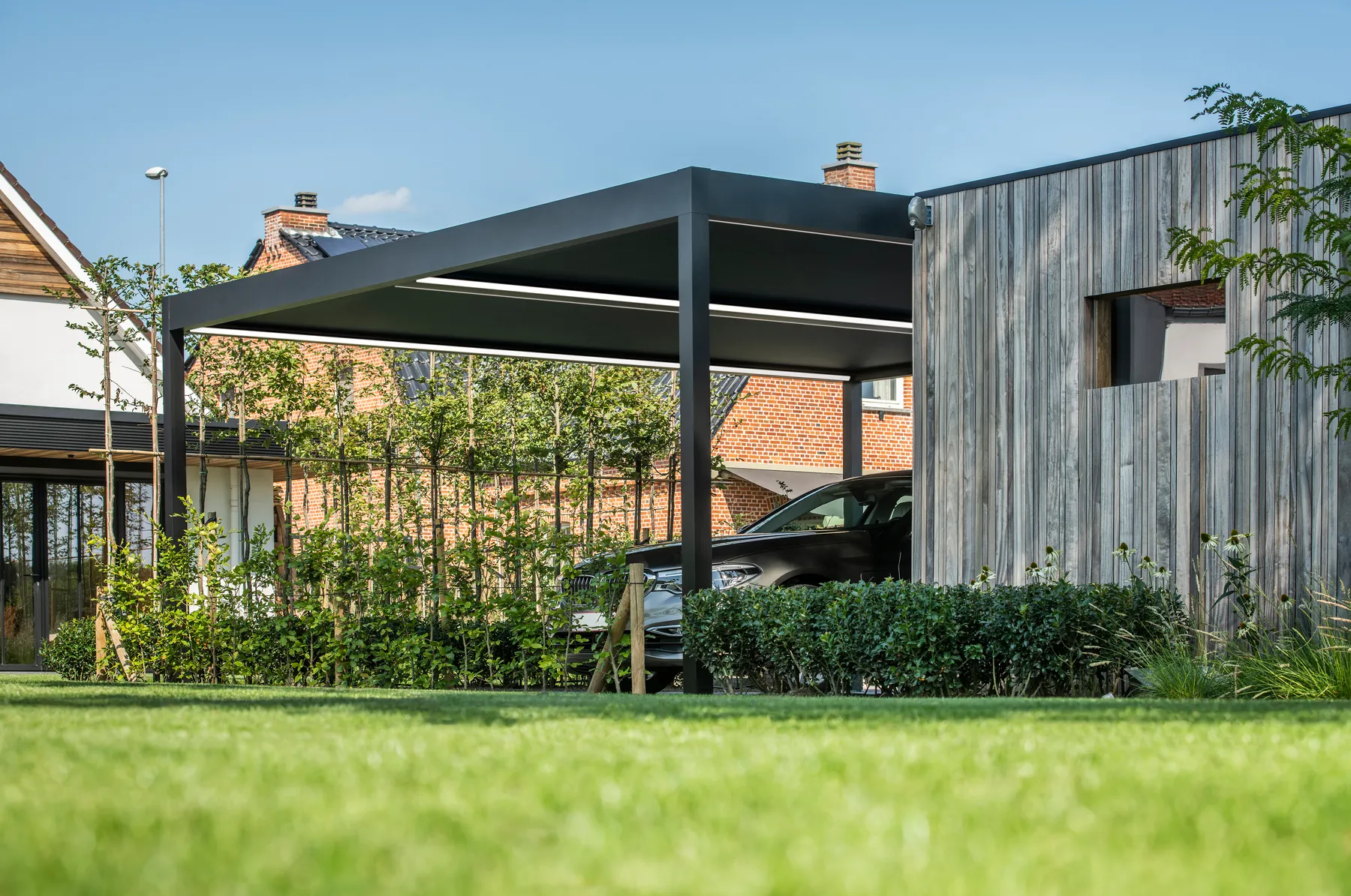 Carport mit Lichtband unter dem Dach, aus der Rasenperspektive fotografiert, dahinter ein Auto und ein Holzhaus