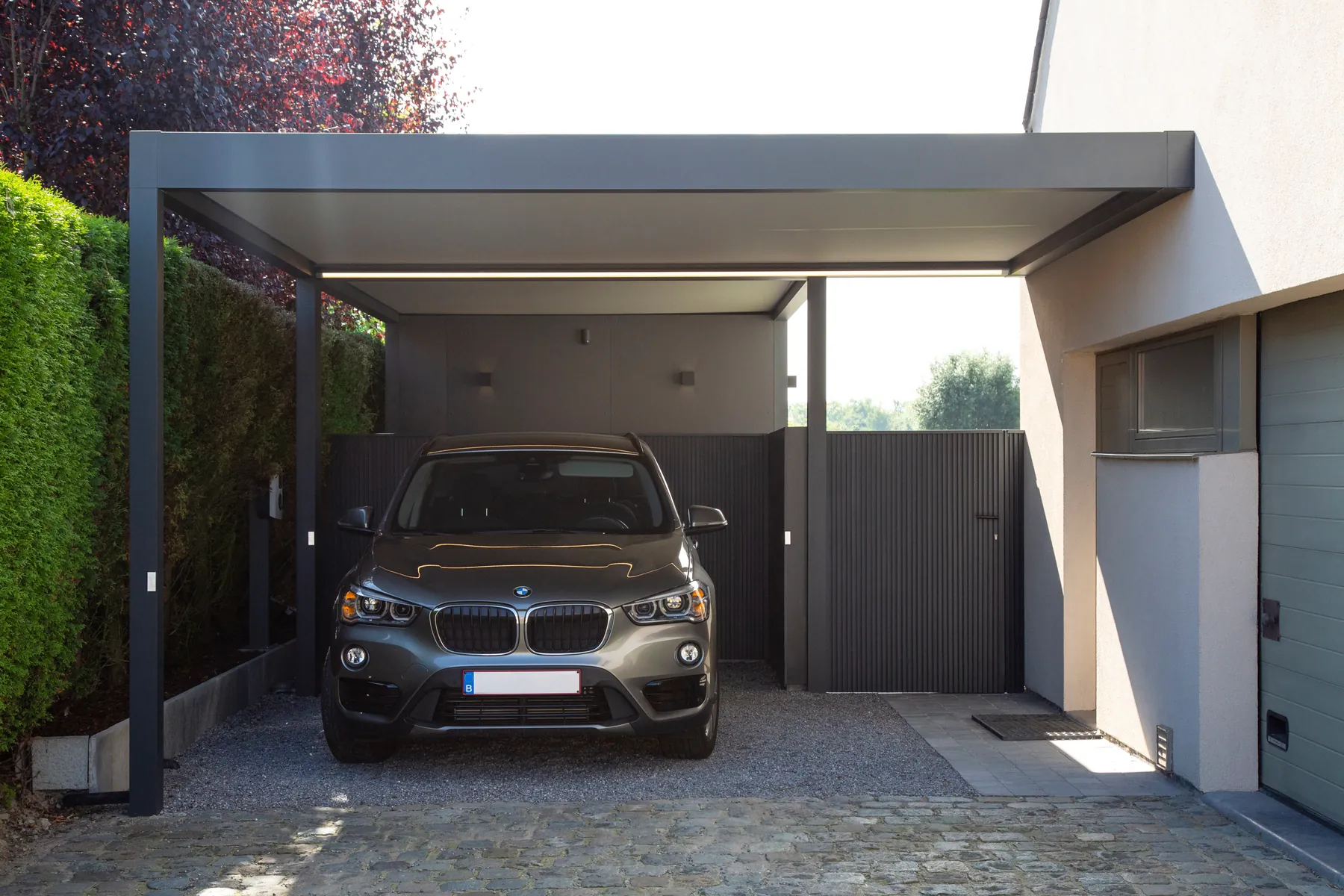 Carport mit Seitenwänden und Lichtband unter dem Dach, darunter ein SUV in Frontansicht, rechts das Wohnhaus