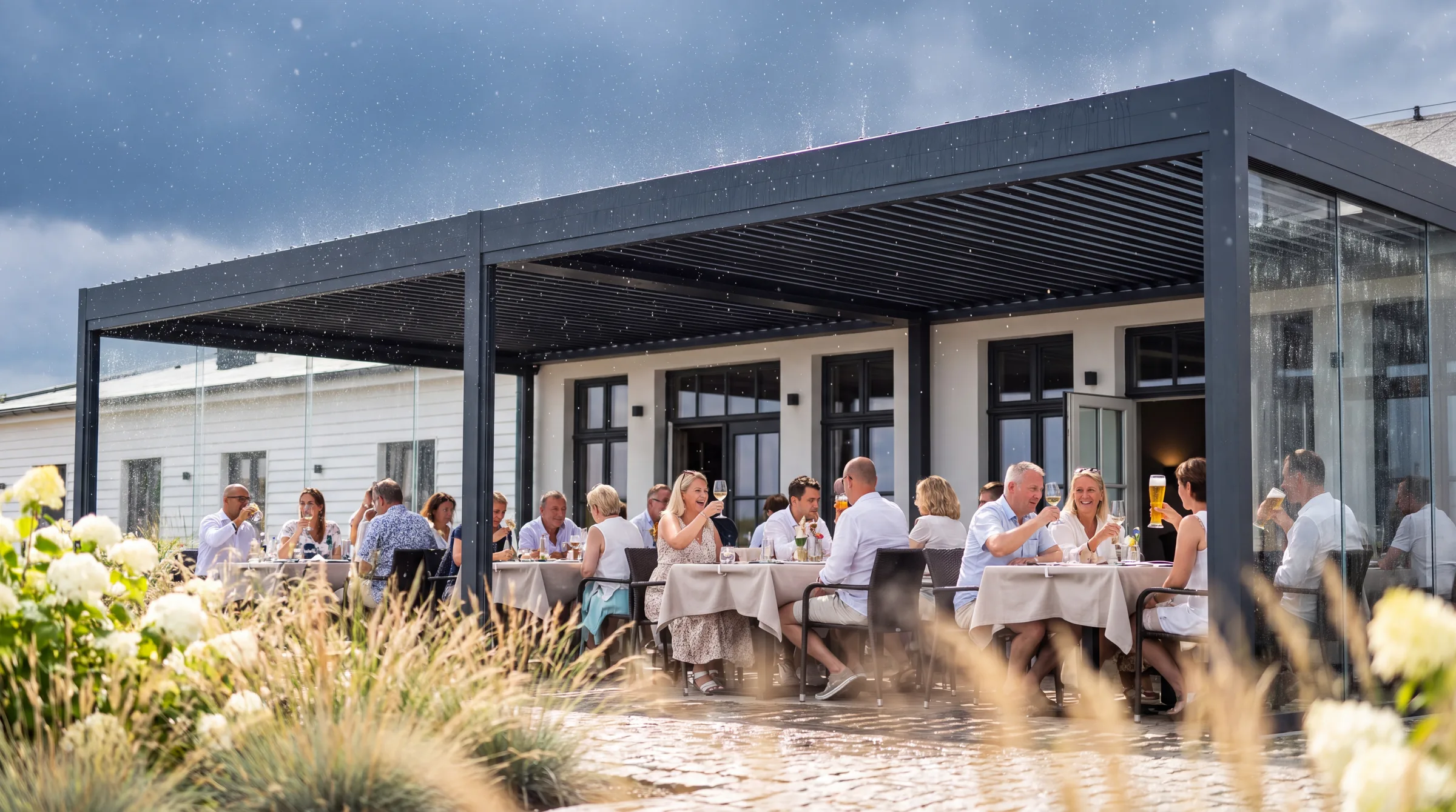 Symbolbild, mit KI erstellt: Überdachte Restaurantterrasse bei Regen, Gäste an gedeckten Tischen unter einem Lamellendach