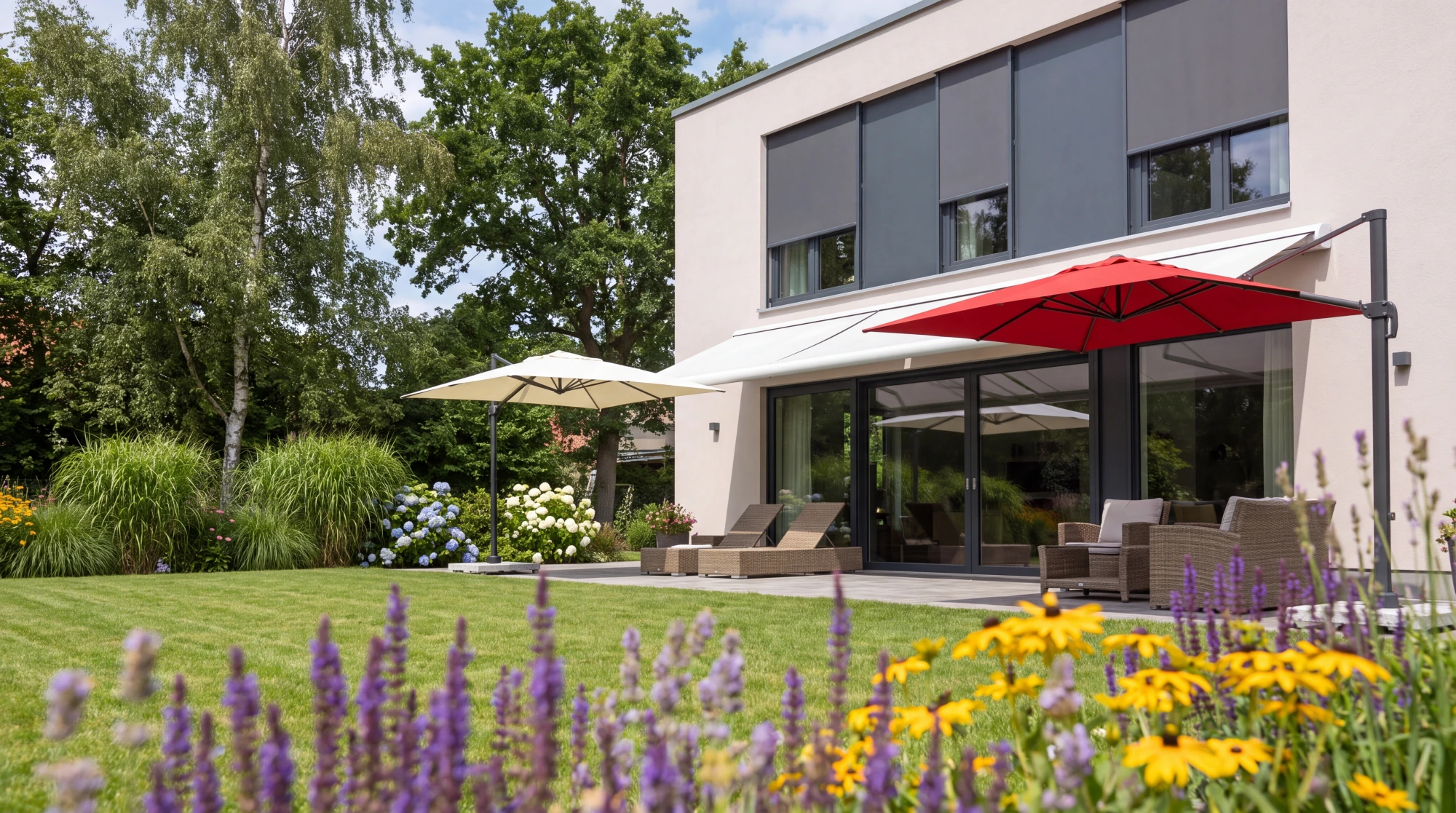 Terrasse mit ausgefahrener Kassettenmarkise und freistehendem rotem Sonnenschirm an einem modernen weißen Haus am Tag, davor ein blühender Garten mit Lavendel